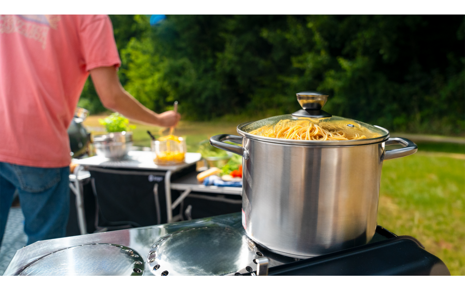 Ein großer Kochtopf auf einem Grill im Freien, im Hintergrund eine Person in einem rosa T-Shirt