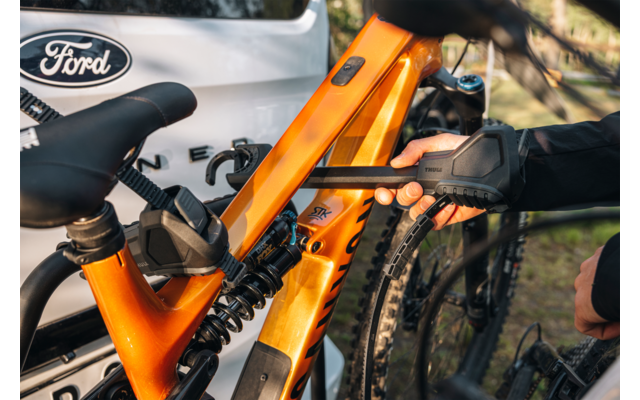 Orangefarbener Mountainbike-Rahmen mit Hinterradfederung, Sattel und grobstolligem Hinterreifen, an einer schwarzen Trägerbefestigung geklemmt; behandschuhte Hand; weißes Auto mit Ford-Logo im Hintergrund