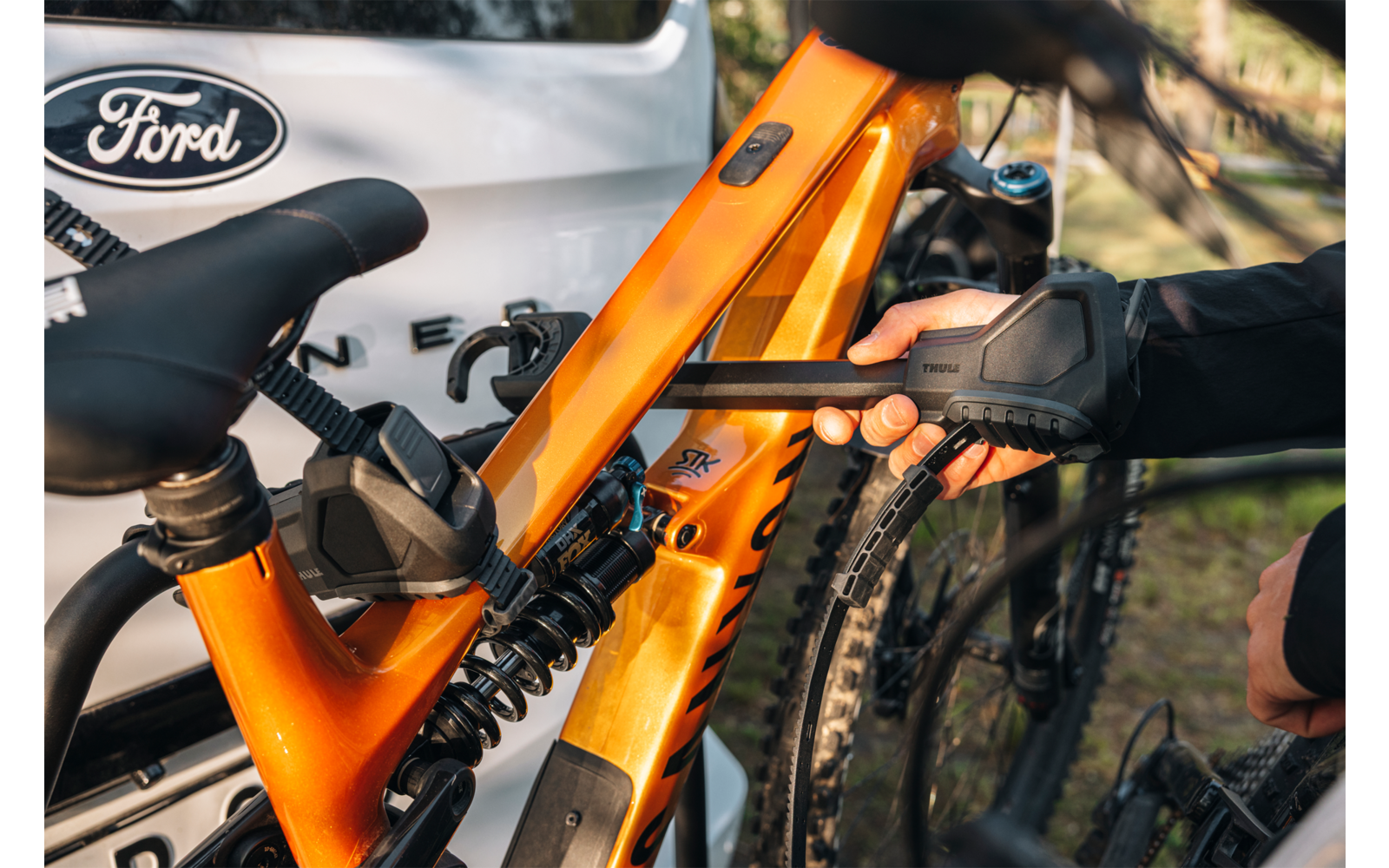 Orangefarbener Mountainbike-Rahmen mit Hinterradfederung, Sattel und grobstolligem Hinterreifen, an einer schwarzen Trägerbefestigung geklemmt; behandschuhte Hand; weißes Auto mit Ford-Logo im Hintergrund