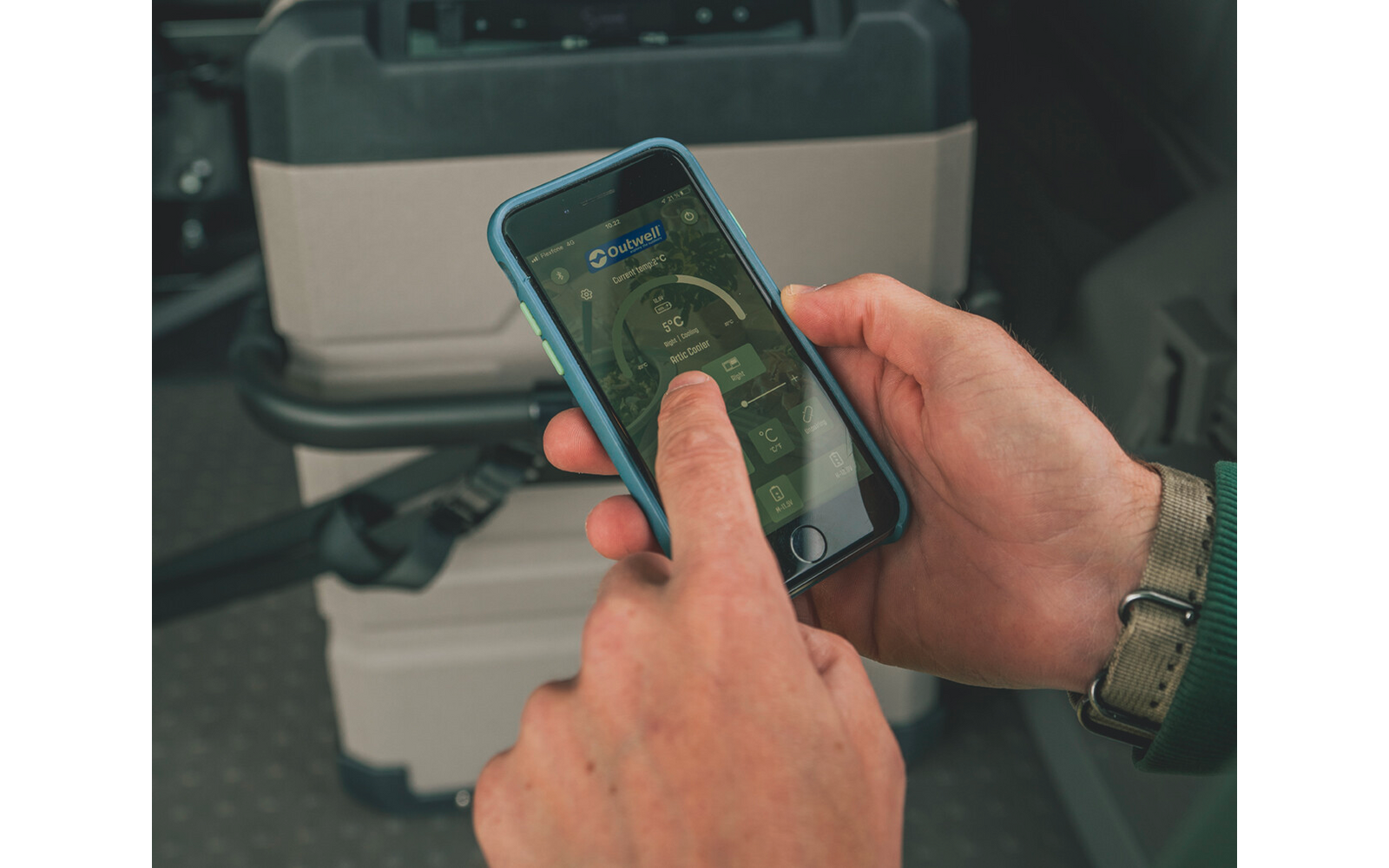 Zwei Hände halten ein Smartphone mit blauer Hülle; ein Daumen berührt den Bildschirm. Im Hintergrund ein grauer Transportkoffer und eine Sitzfläche