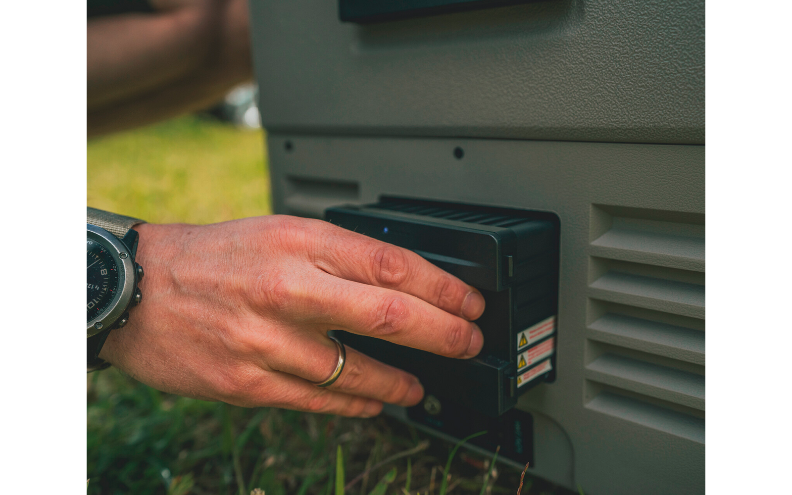 Nahaufnahme von Händen, die ein schwarzes Akkupack in ein graues Gerät einsetzen; Gras im Hintergrund; sichtbarer Ring und Armbanduhr