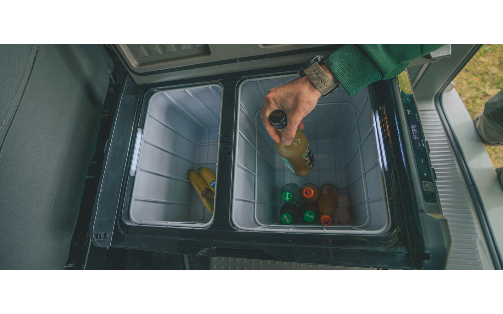 Hand mit Armbanduhr und grünem Ärmel legt eine Glasflasche in eine geöffnete Kühlbox mit zwei Fächern, darin weitere Flaschen und Dosen