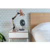 Table de chevet blanche avec tiroir, lampe cuivrée et petit ventilateur noir ; à droite un lit en bois avec couette blanche ; papier peint géométrique bleu‑gris ; à gauche une feuille de plante