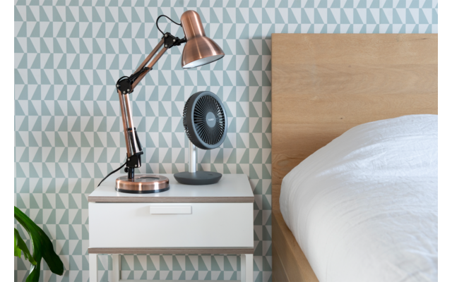 Table de chevet blanche avec tiroir, lampe cuivrée et petit ventilateur noir ; à droite un lit en bois avec couette blanche ; papier peint géométrique bleu‑gris ; à gauche une feuille de plante