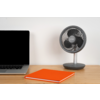 Partie d’un ordinateur portable avec écran noir à gauche, un carnet orange fermé au centre et un ventilateur de table gris à droite sur une surface en bois devant un mur blanc