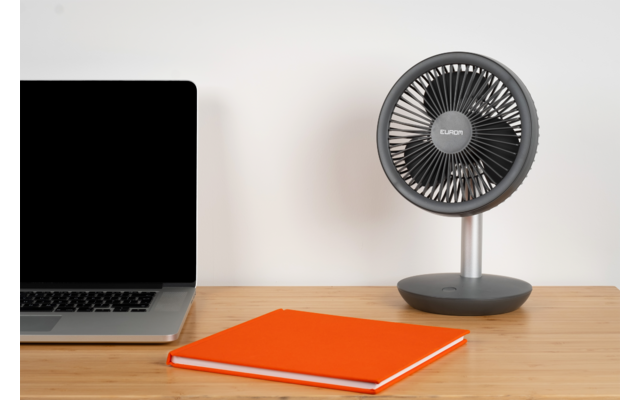 Partie d’un ordinateur portable avec écran noir à gauche, un carnet orange fermé au centre et un ventilateur de table gris à droite sur une surface en bois devant un mur blanc