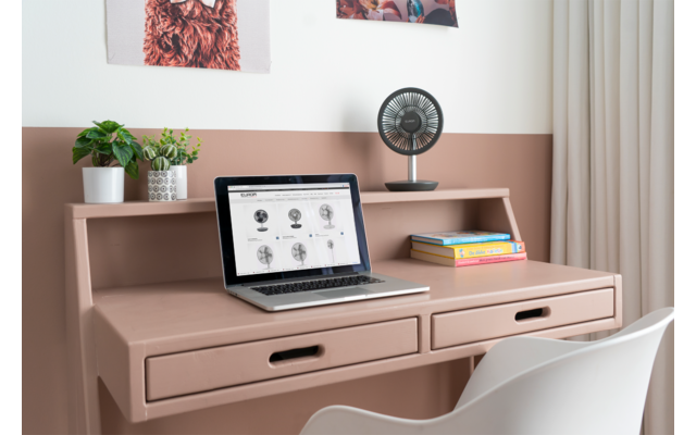 Bureau rose avec meuble de rangement et deux tiroirs. Sur le plateau : ordinateur portable ouvert, petit ventilateur, deux livres et une plante en pot. Dossier de chaise blanc au premier plan, deux tableaux au mur, mur bicolore jusqu'à mi-hauteur et rideau
