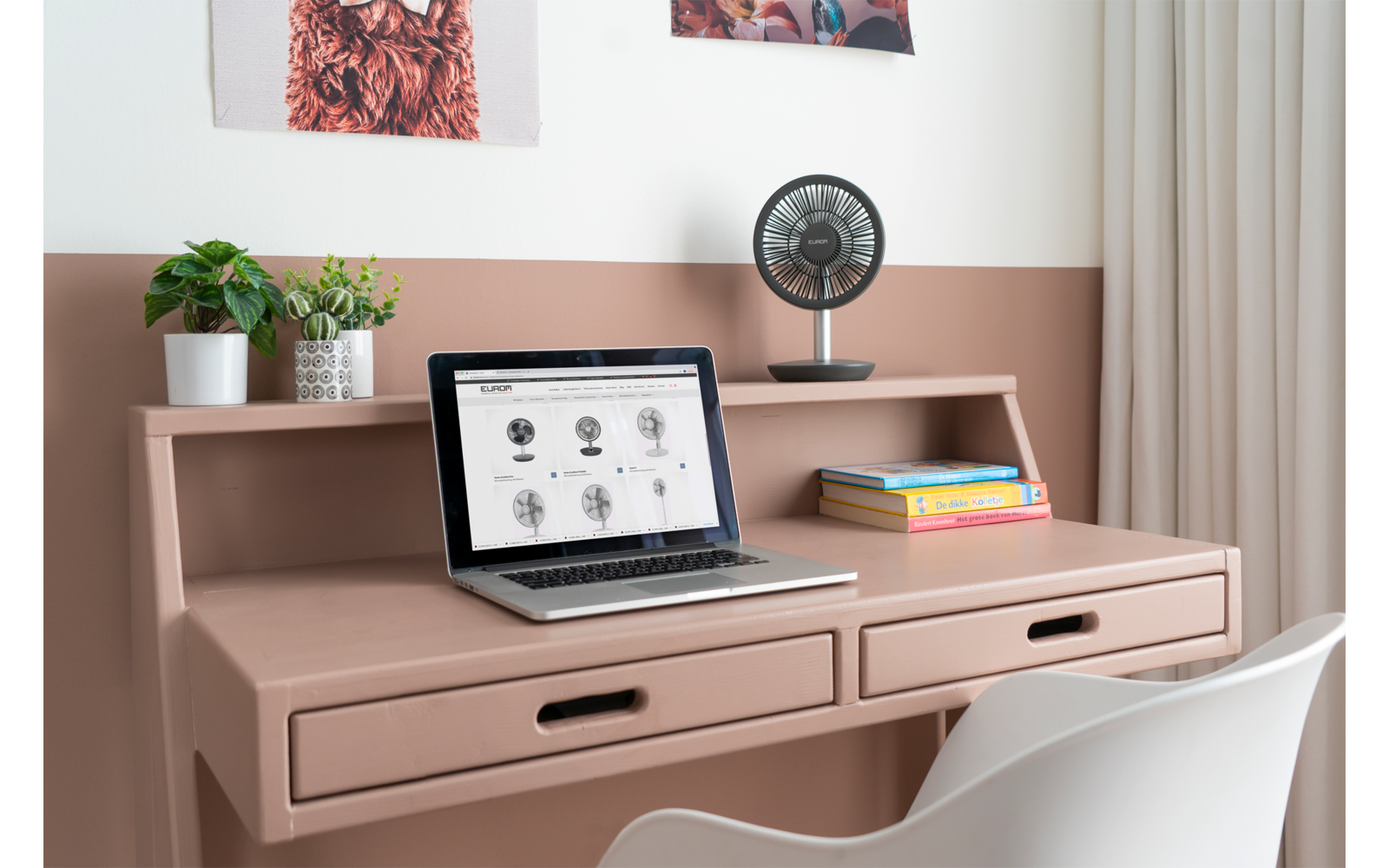 Bureau rose avec meuble de rangement et deux tiroirs. Sur le plateau : ordinateur portable ouvert, petit ventilateur, deux livres et une plante en pot. Dossier de chaise blanc au premier plan, deux tableaux au mur, mur bicolore jusqu'à mi-hauteur et rideau