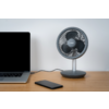 Sur un bureau en bois, à gauche un ordinateur portable ouvert avec écran noir, à droite un petit ventilateur de table rond et devant un smartphone noir ; un câble relie le ventilateur à l'ordinateur