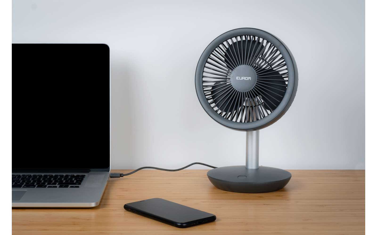 Sur un bureau en bois, à gauche un ordinateur portable ouvert avec écran noir, à droite un petit ventilateur de table rond et devant un smartphone noir ; un câble relie le ventilateur à l'ordinateur