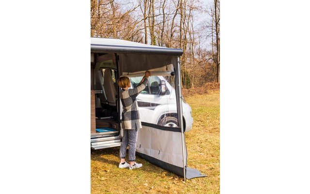 Person neben Wohnmobil befestigt seitlichen Wetterschutz/Markisenverkleidung, grasbewachsene Fläche und kahle Bäume im Hintergrund