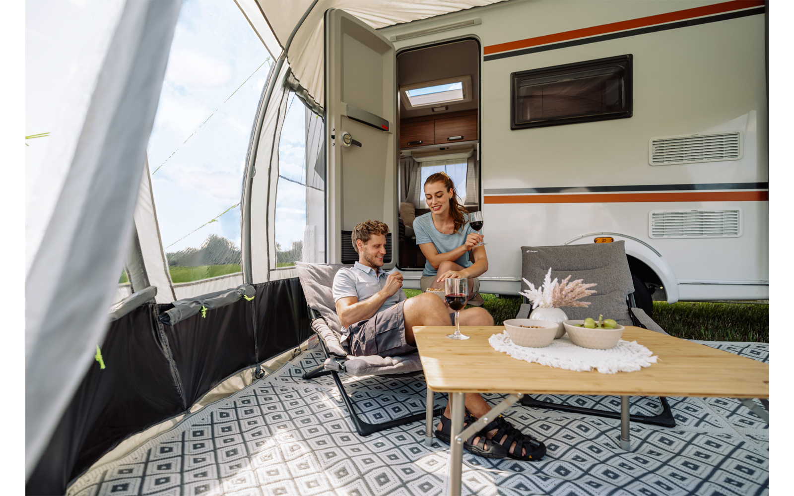 Vor einem geöffneten Wohnmobil: ein mit Teppich ausgelegtes Vorzelt, ein Mann sitzt in einer Campingliege, eine Frau gießt Flüssigkeit in eine Tasse auf einem Holztisch mit Schale und Vase; im Hintergrund Wiese und Himmel