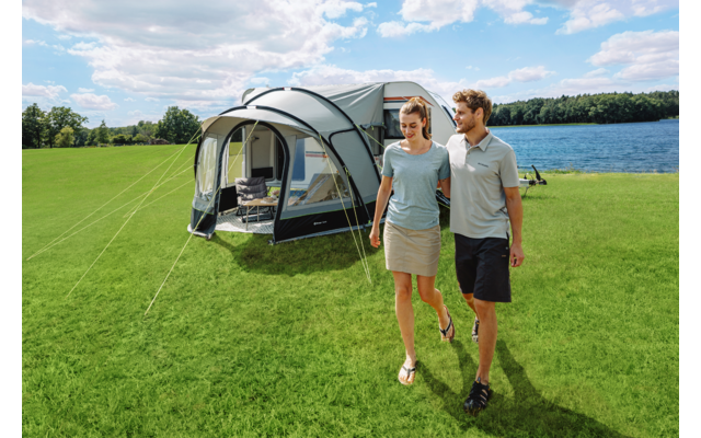 Ein Mann und eine Frau gehen auf einer grünen Wiese; hinter ihnen steht ein großes Zelt mit Vorbau, im Hintergrund ein See, Bäume und ein wolkiger Himmel