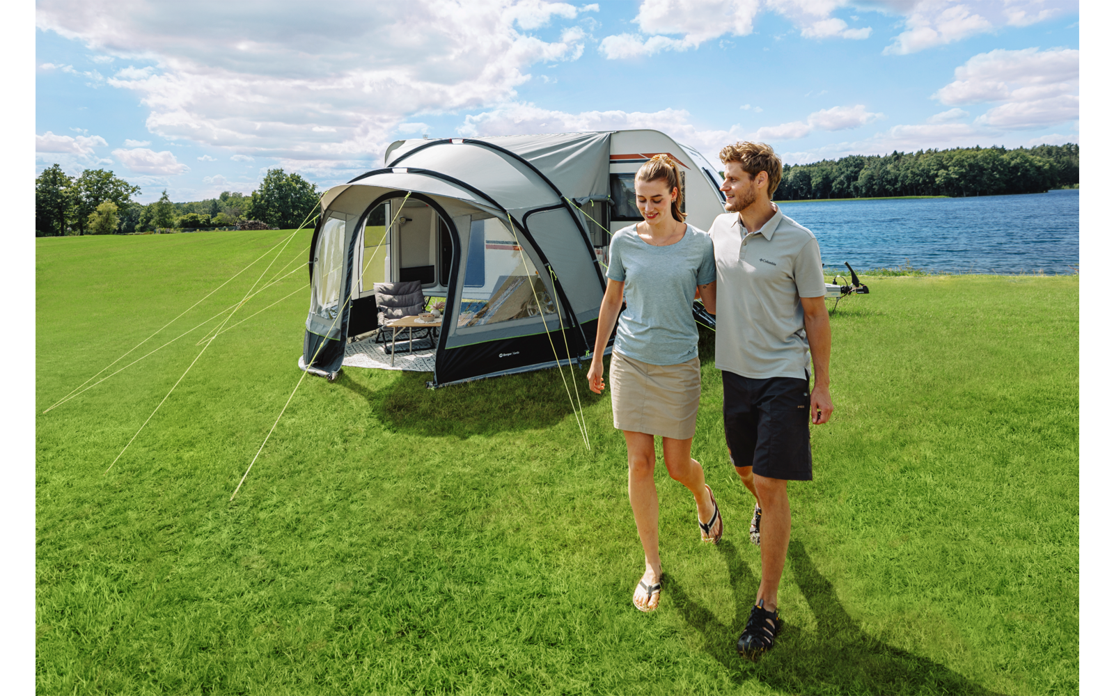 Ein Mann und eine Frau gehen auf einer grünen Wiese; hinter ihnen steht ein großes Zelt mit Vorbau, im Hintergrund ein See, Bäume und ein wolkiger Himmel