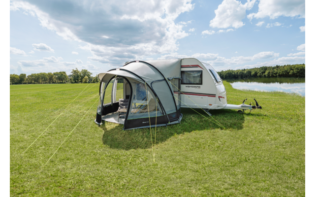 Weißer Wohnwagen mit grauem aufblasbarem Vorzelt auf grüner Wiese neben einem See, teilweise bewölkter Himmel