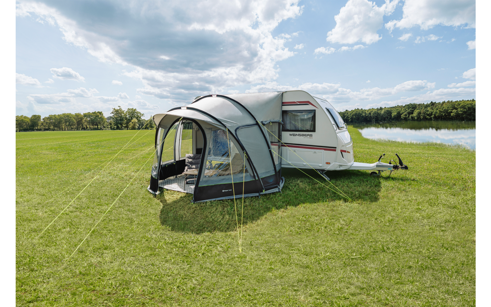 Weißer Wohnwagen mit grauem aufblasbarem Vorzelt auf grüner Wiese neben einem See, teilweise bewölkter Himmel