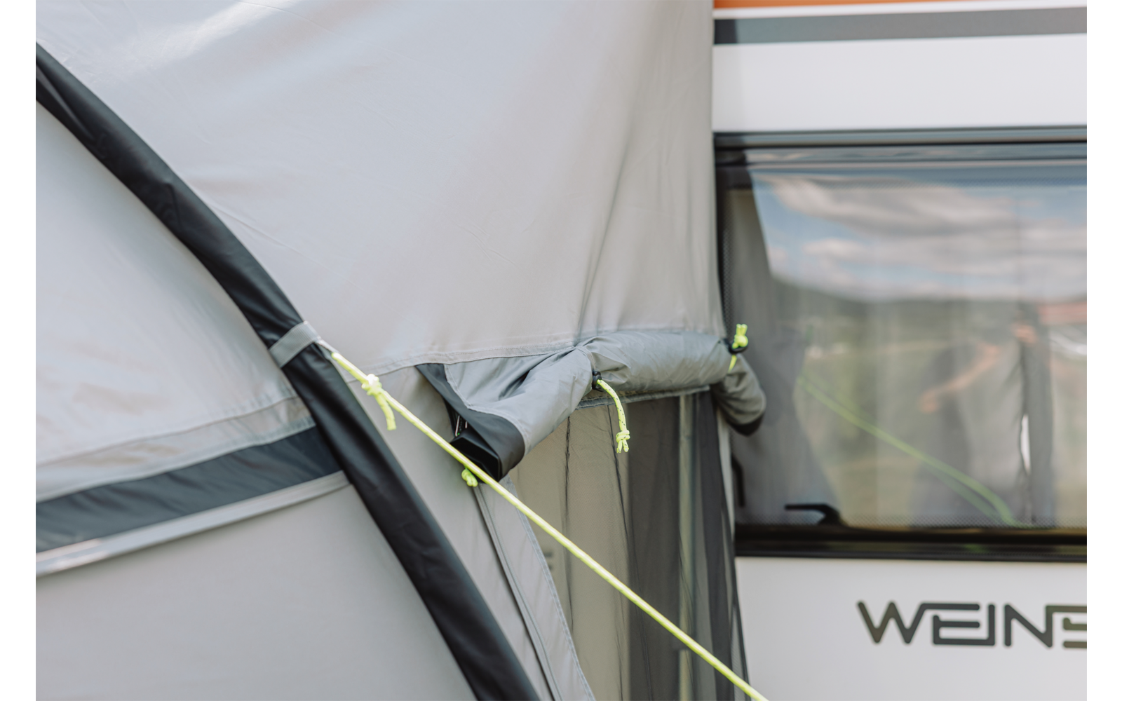 Nahaufnahme eines grauen Vorzelts mit schwarzem Gestängebogen, neongelber Abspannleine und einem angrenzenden weißen Wohnwagen mit Fenster