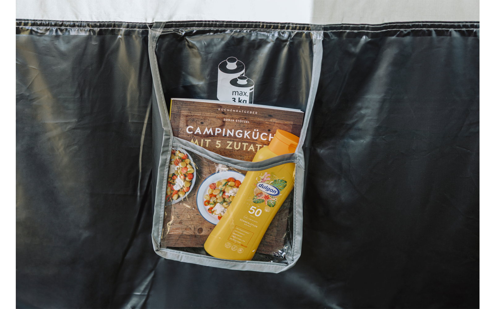 Transparentes, hängendes Fach mit einer gelben Sonnencreme‑Flasche (SPF 50), einer braunen Verpackung mit der Aufschrift Campingküche mit 5 Zutaten und einer kleinen Dose oben