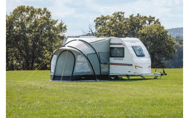 Weißer Wohnwagen mit angebautem Vorzelt auf grünem Feld, Bäume und teils bewölkter Himmel im Hintergrund