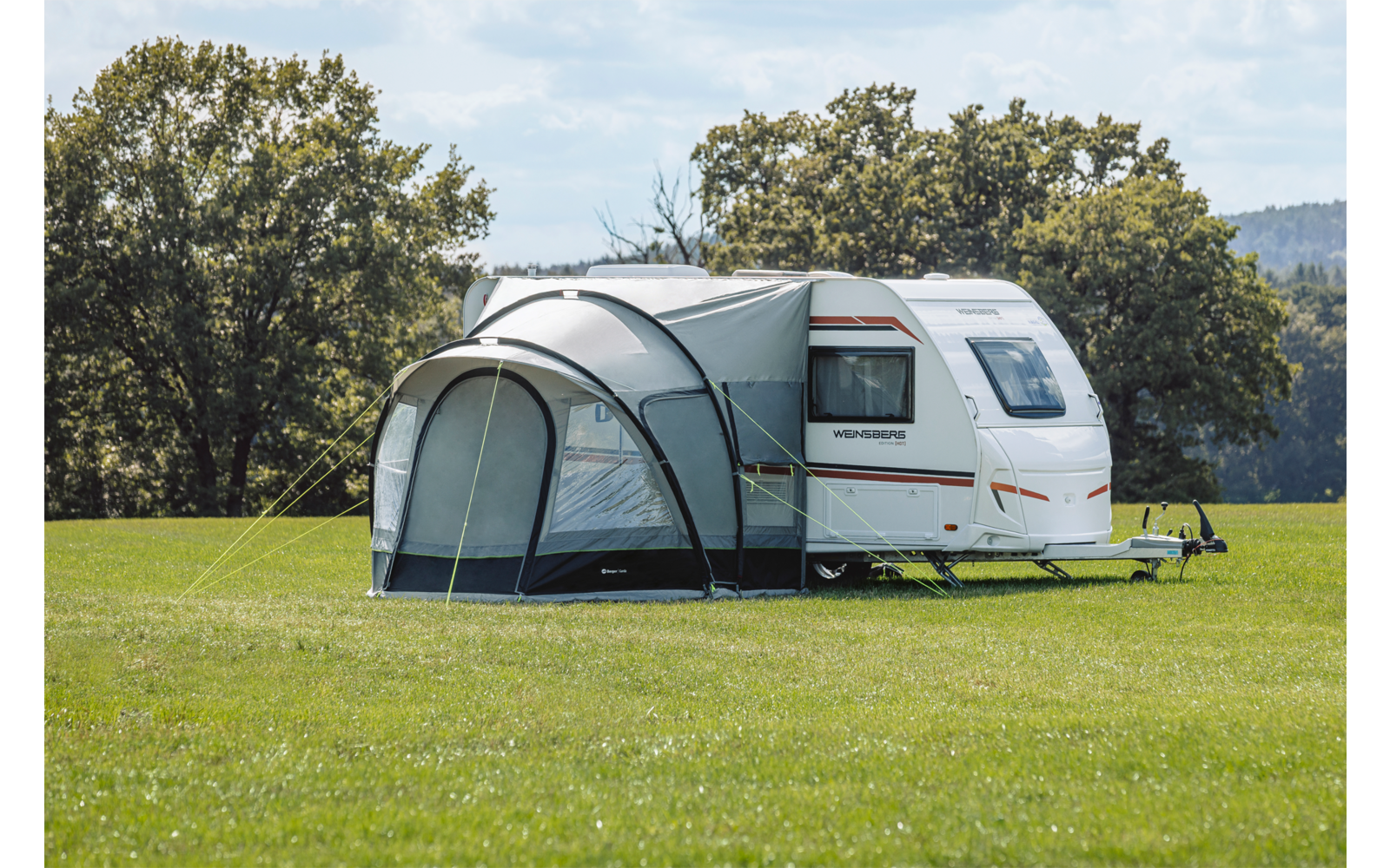 Weißer Wohnwagen mit angebautem Vorzelt auf grünem Feld, Bäume und teils bewölkter Himmel im Hintergrund