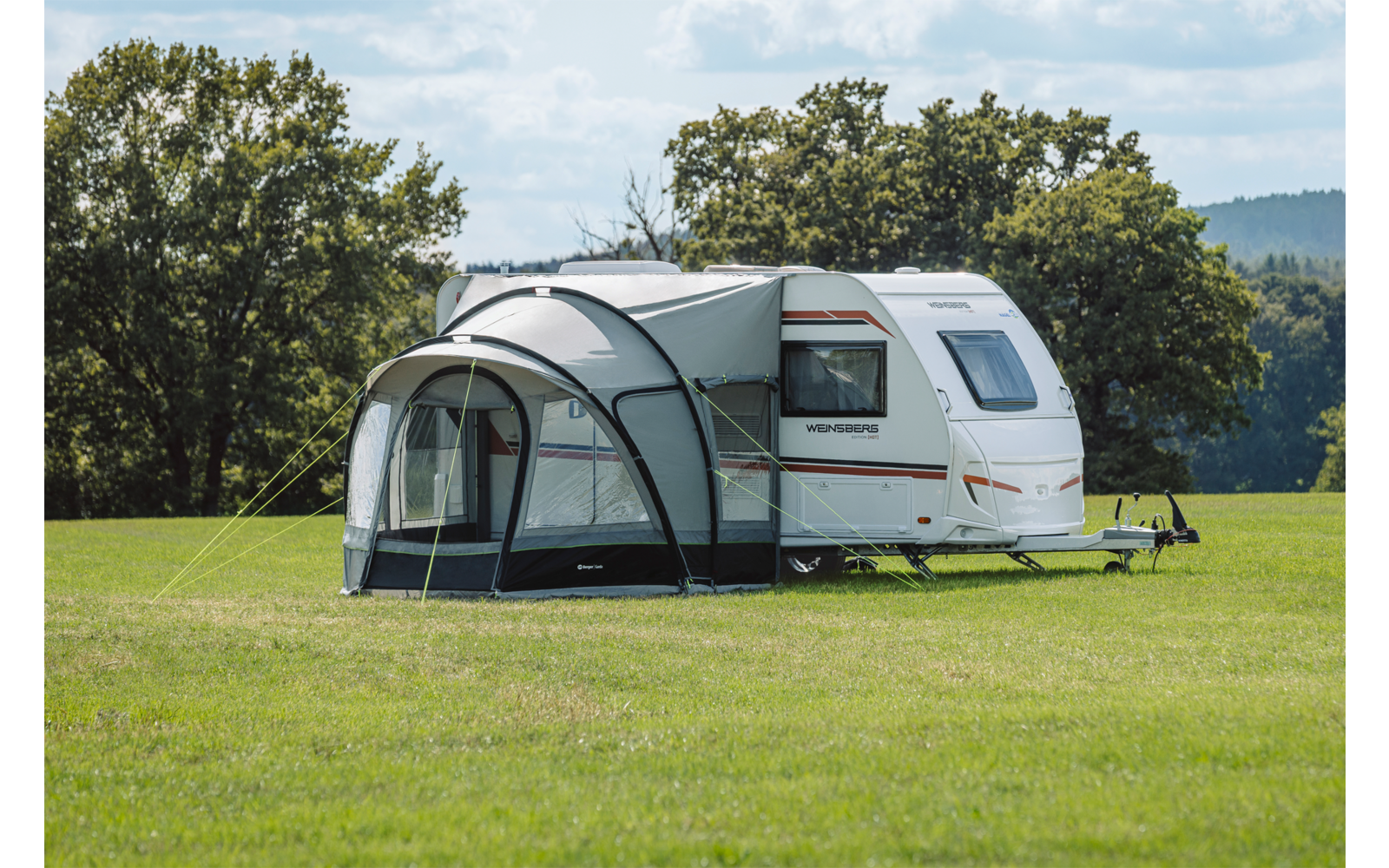 Weißer Wohnwagen mit grauem Vorzelt auf einer grünen Wiese, Bäume und bewölkter Himmel im Hintergrund