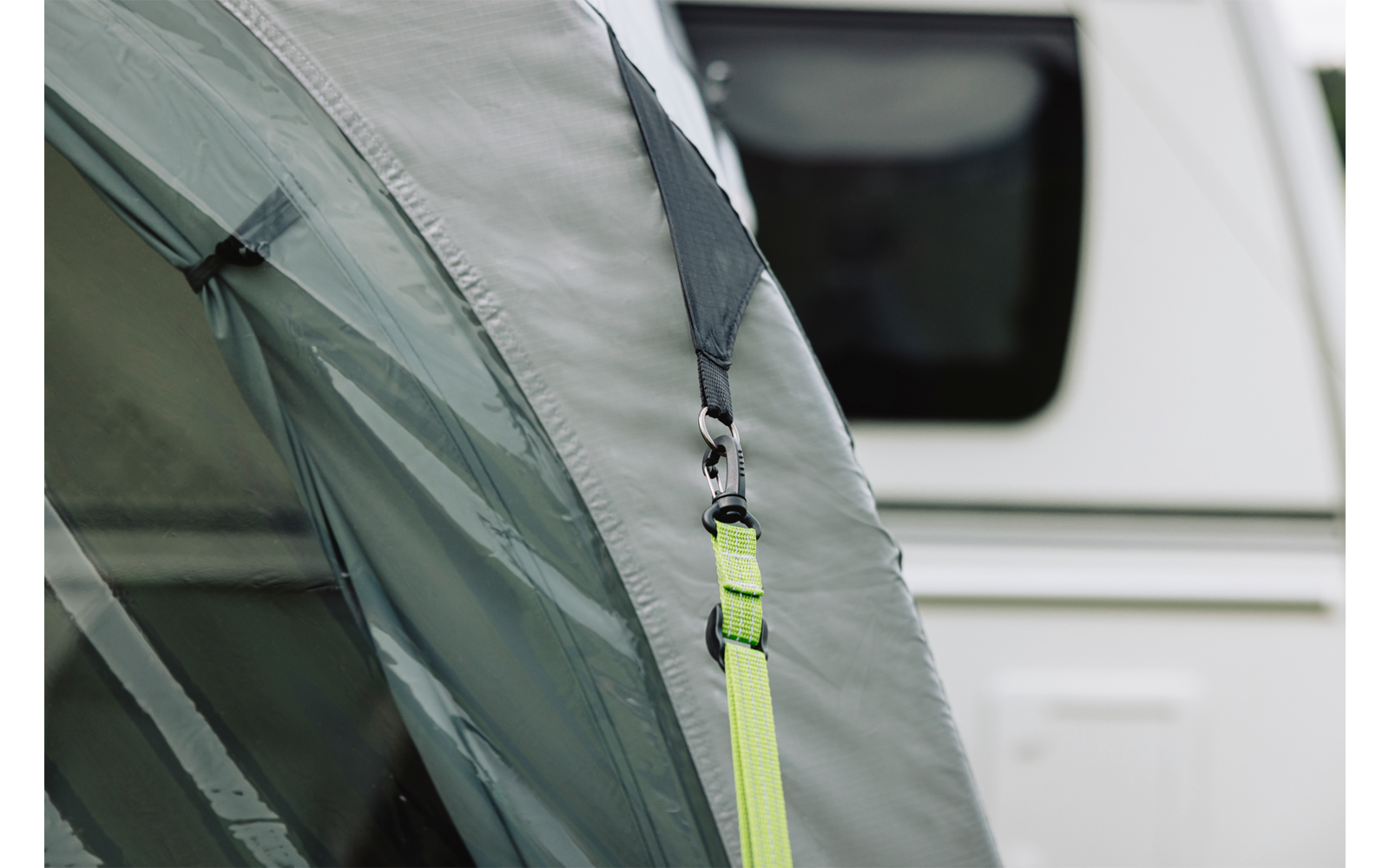 Grauer Zeltstoff mit schwarzem Gurt, Metallkarabiner und neongrünem Spanngurt; im Hintergrund weißer Wohnwagen mit Fenster