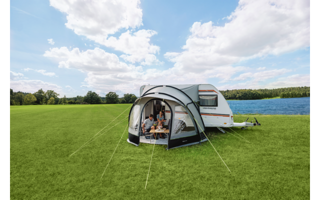Wohnwagen mit angebautem Vorzelt auf grüner Wiese neben einem See, drei Personen sitzen im Vorzelt