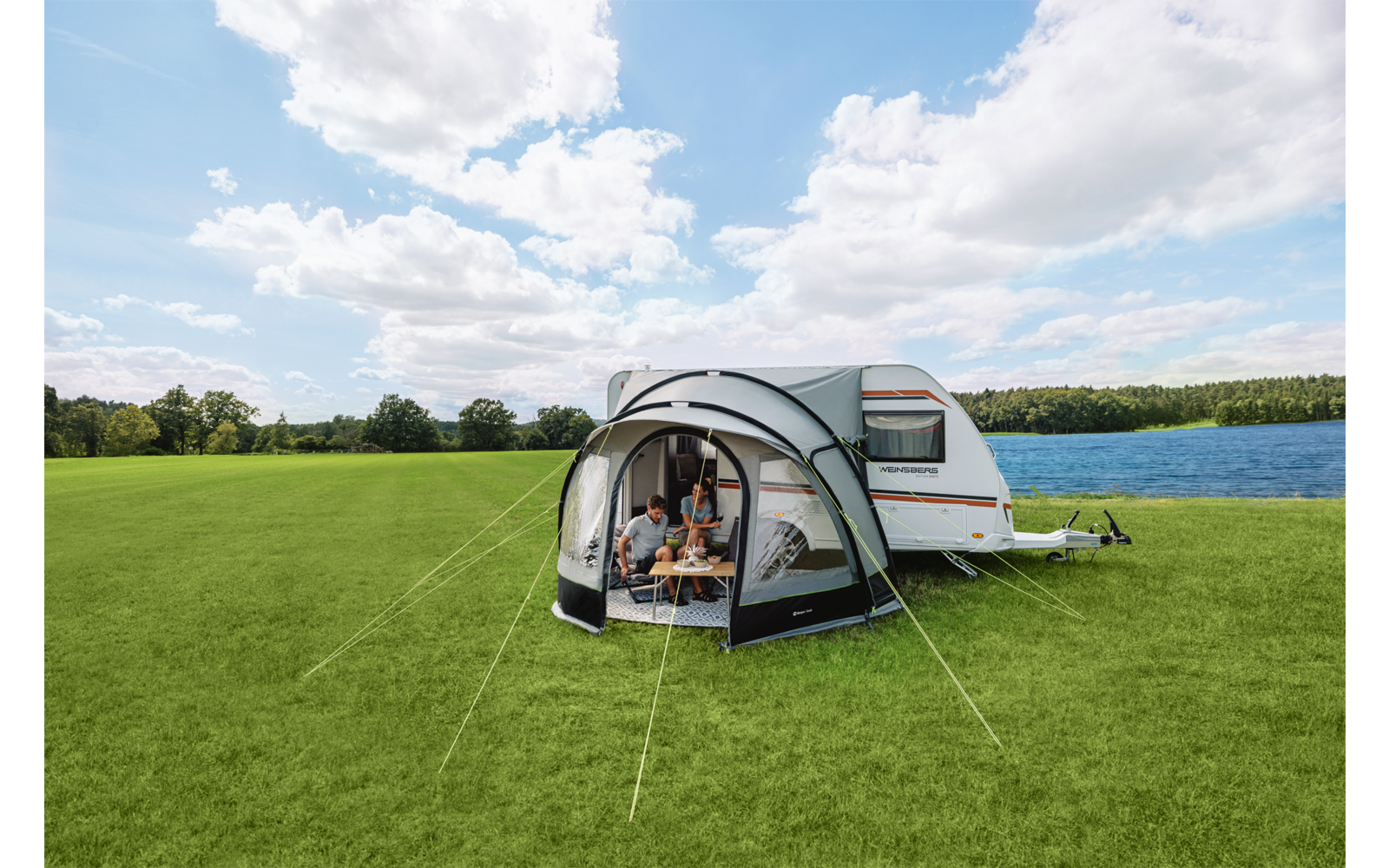 Wohnwagen mit angebautem Vorzelt auf grüner Wiese neben einem See, drei Personen sitzen im Vorzelt