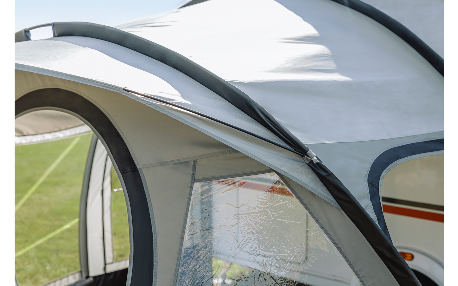 Weiße Plane mit geschwungenen dunkelgrauen Stangen, durchsichtig strukturiertes Fenster, seitlich sichtbare weiße Fahrzeugwand mit orangefarbenem Streifen, grüner Rasen im Hintergrund