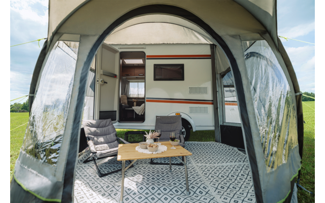 Offenes Vorzelt an einem Wohnwagen auf einer Wiese, darin zwei Klappstühle und ein niedriger Tisch mit Teekanne, Tassen und Snacks auf einem gemusterten Bodenbelag