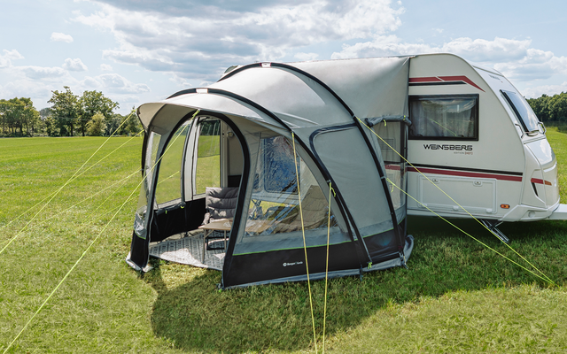 Weißer Wohnwagen mit grauem aufblasbarem Vorzelt auf grüner Wiese neben einem See, teilweise bewölkter Himmel