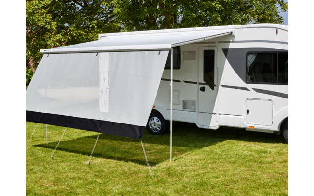 Camper bianco con tendalino aperto e schermo laterale su prato, alberi sullo sfondo