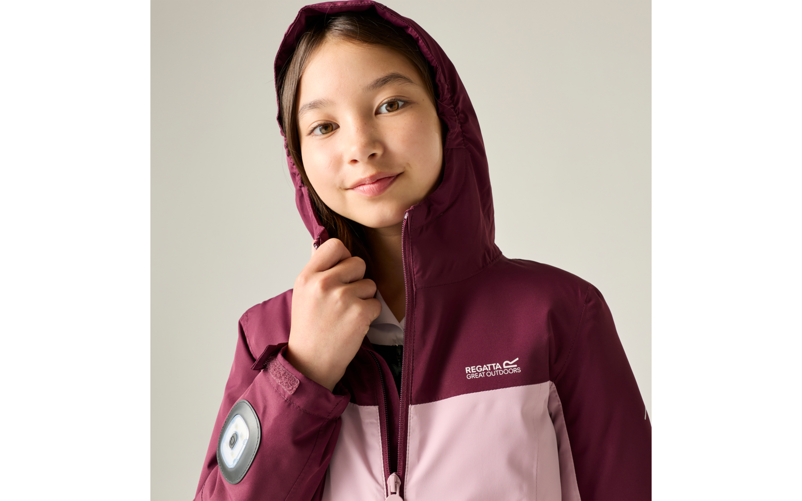 Fille portant une veste à capuche bordeaux et rose clair, capuche portée, tenant la fermeture éclair, logo de marque visible sur la poitrine, écusson rond sur la manche gauche