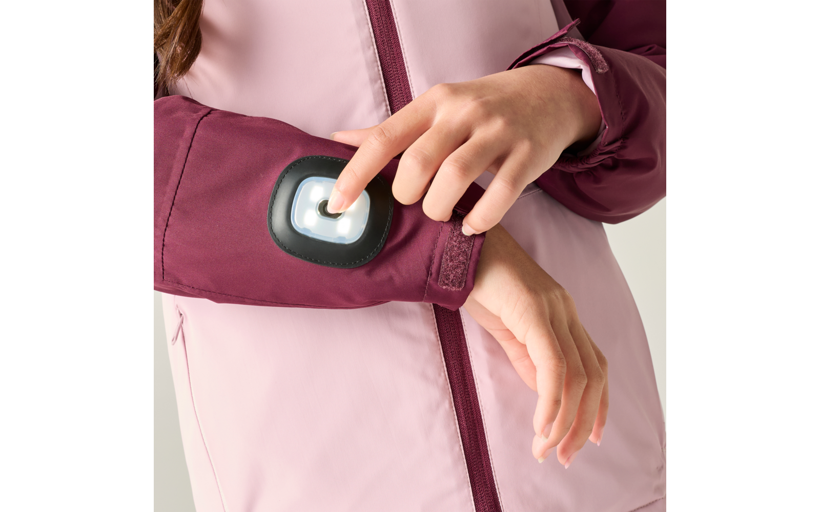 Gros plan de mains appuyant sur un module lumineux blanc dans un boîtier noir sur la manche d'une veste rose clair et bordeaux; fermeture éclair et velcro visibles
