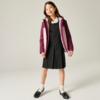 Fille debout sur fond clair, portant une robe noire plissée, une blouse blanche, une veste à capuche bordeaux avec doublure plus claire, chaussettes blanches chevillères et chaussures noires, léger sourire