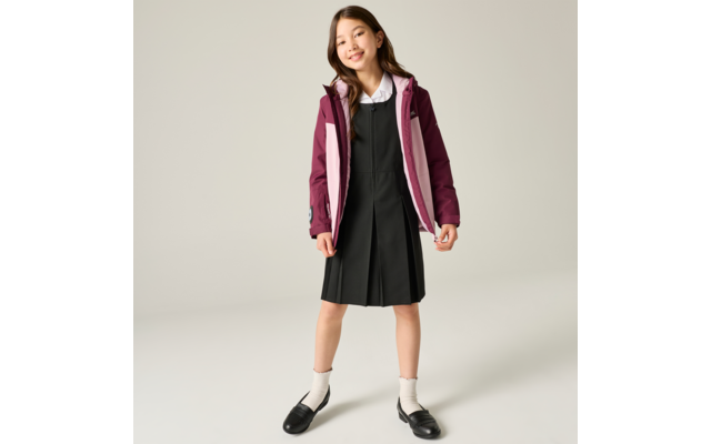 Fille debout sur fond clair, portant une robe noire plissée, une blouse blanche, une veste à capuche bordeaux avec doublure plus claire, chaussettes blanches chevillères et chaussures noires, léger sourire