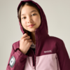 Fille portant une veste à capuche bordeaux et rose clair, capuche portée, tenant la fermeture éclair, logo de marque visible sur la poitrine, écusson rond sur la manche gauche