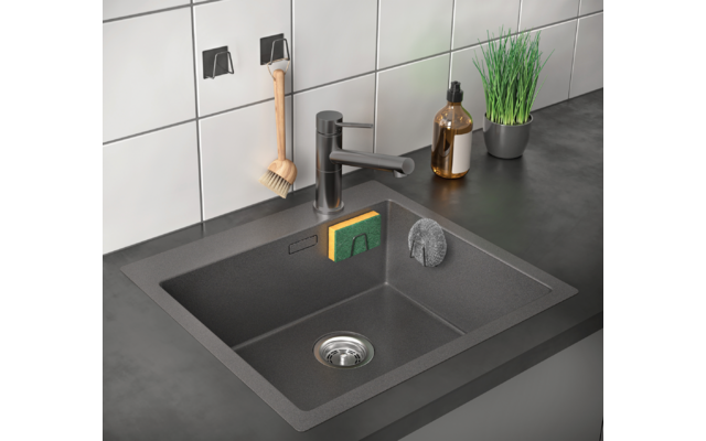 Schwarze Küchenspüle mit Wasserhahn, daneben Seifenspender und Pflanze auf grauer Arbeitsplatte