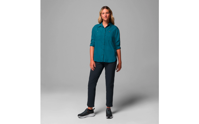 Femme debout devant un fond gris, porte une chemise bleue et un pantalon noir