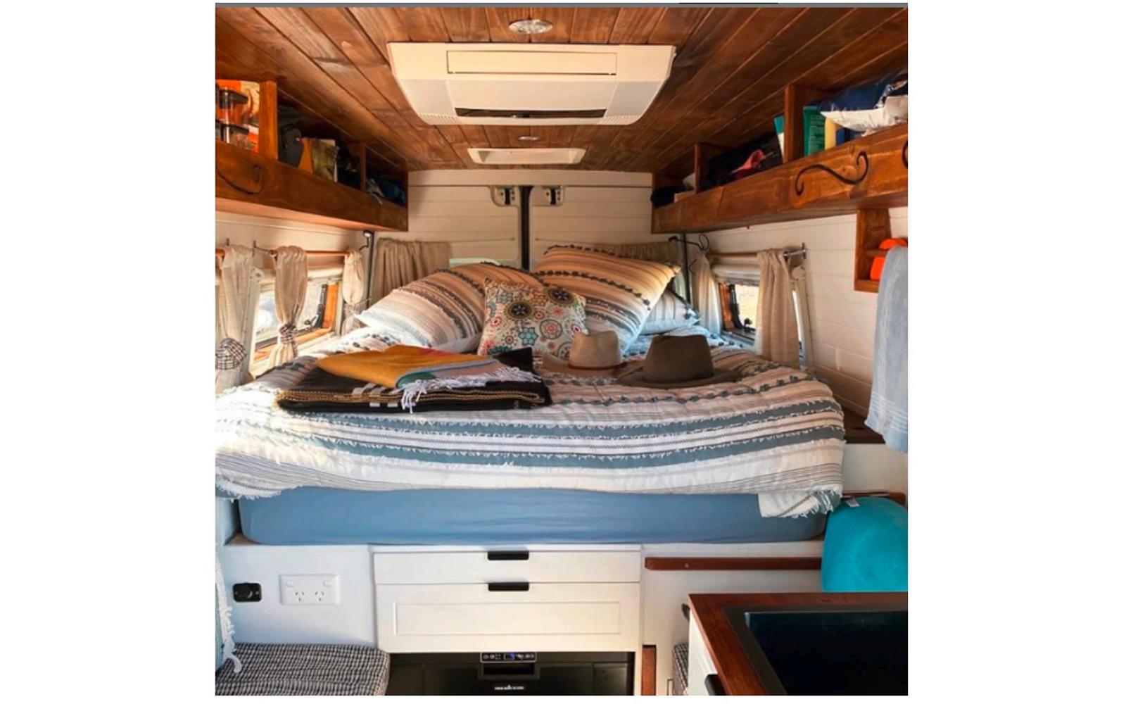 Bed met gemêleerd beddengoed en kussens, twee hoeden en gevouwen textiel op het bed, houten plafond met ingebouwde airco, open planken met spullen, kleine ramen met gordijnen, witte laden en een werkblad met gootsteen
