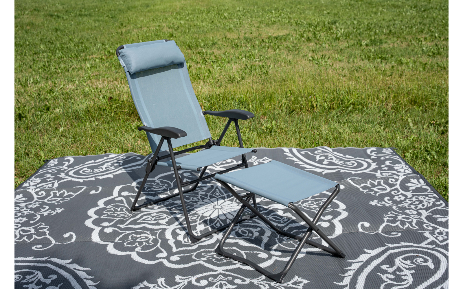 Foldable reclining chair with headrest and matching footstool on a dark gray rug with white floral pattern on green grass