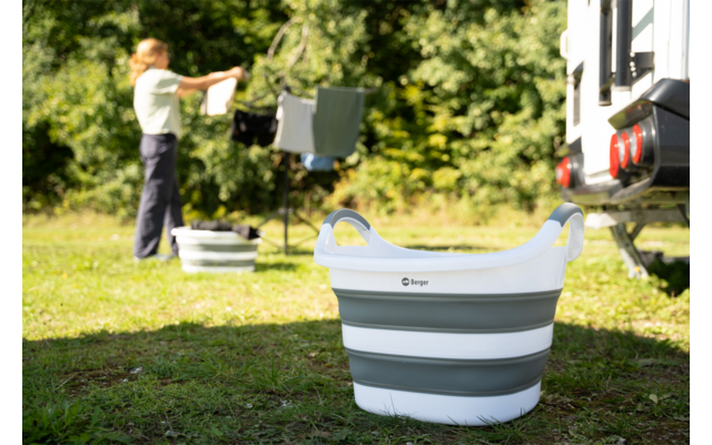 Zusammenfaltbarer weiß-grauer Wäschekorb auf Gras; im Hintergrund hängt eine Person Wäsche neben einem Wohnmobil, Bäume