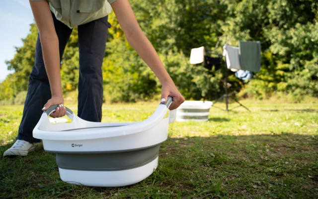 Person von der Taille abwärts, die eine faltbare weiße-graue Wanne mit Griffen auf einer Wiese hält; im Hintergrund Bäume und zwei Wäscheständer