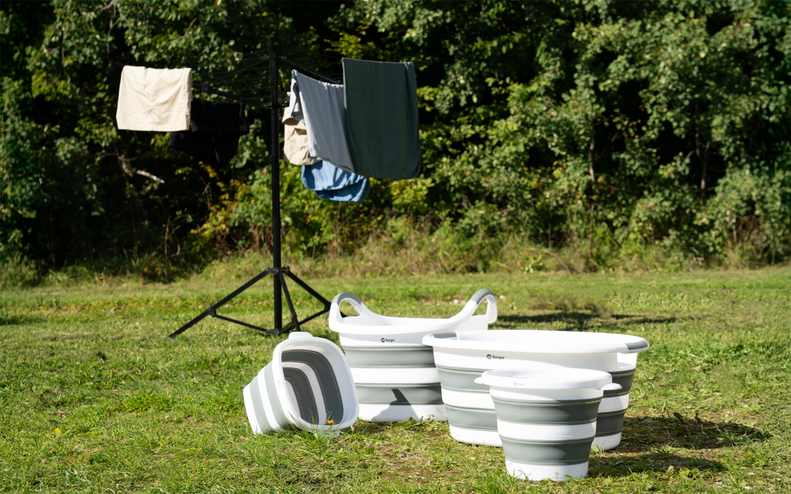 Drei weiß-grau gestreifte, zusammengefaltete Kunststoffwannen auf einer Wiese, eine liegt auf der Seite; im Hintergrund ein Wäscheständer mit hängender Kleidung und Bäume