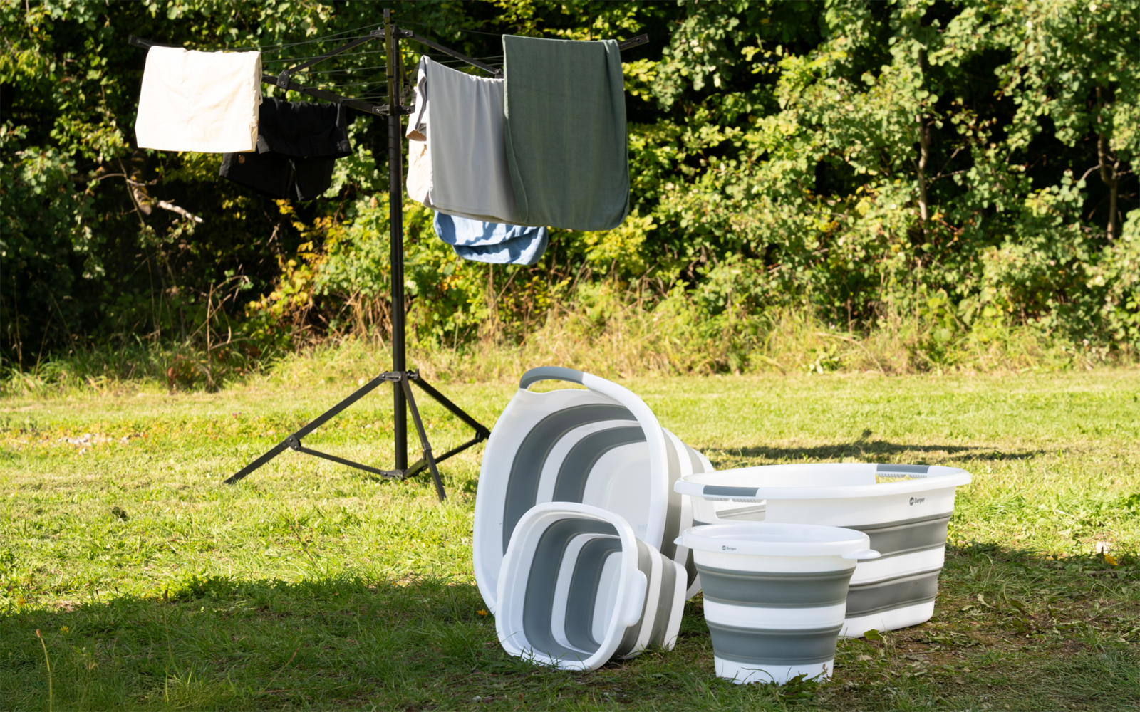 Gestapelte weiße und graue, faltbare Wäschebehälter auf Gras; im Hintergrund eine Wäscheleine an einem Dreibein-Ständer mit aufgehängten Kleidungsstücken und Bäumen