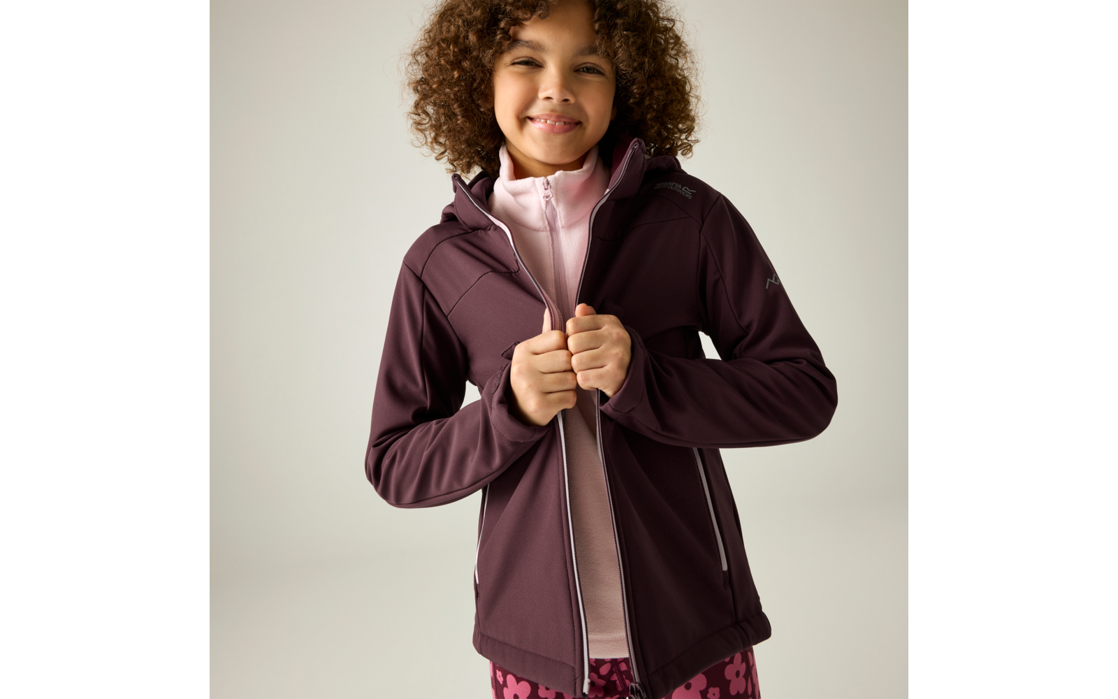 Kind mit lockigem Haar, das eine dunkellila Jacke mit Reißverschluss über einem rosa Oberteil hochzieht, neutraler Hintergrund