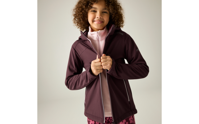 Kind mit lockigem Haar, das eine dunkellila Jacke mit Reißverschluss über einem rosa Oberteil hochzieht, neutraler Hintergrund