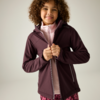 Kind mit lockigem Haar, das eine dunkellila Jacke mit Reißverschluss über einem rosa Oberteil hochzieht, neutraler Hintergrund