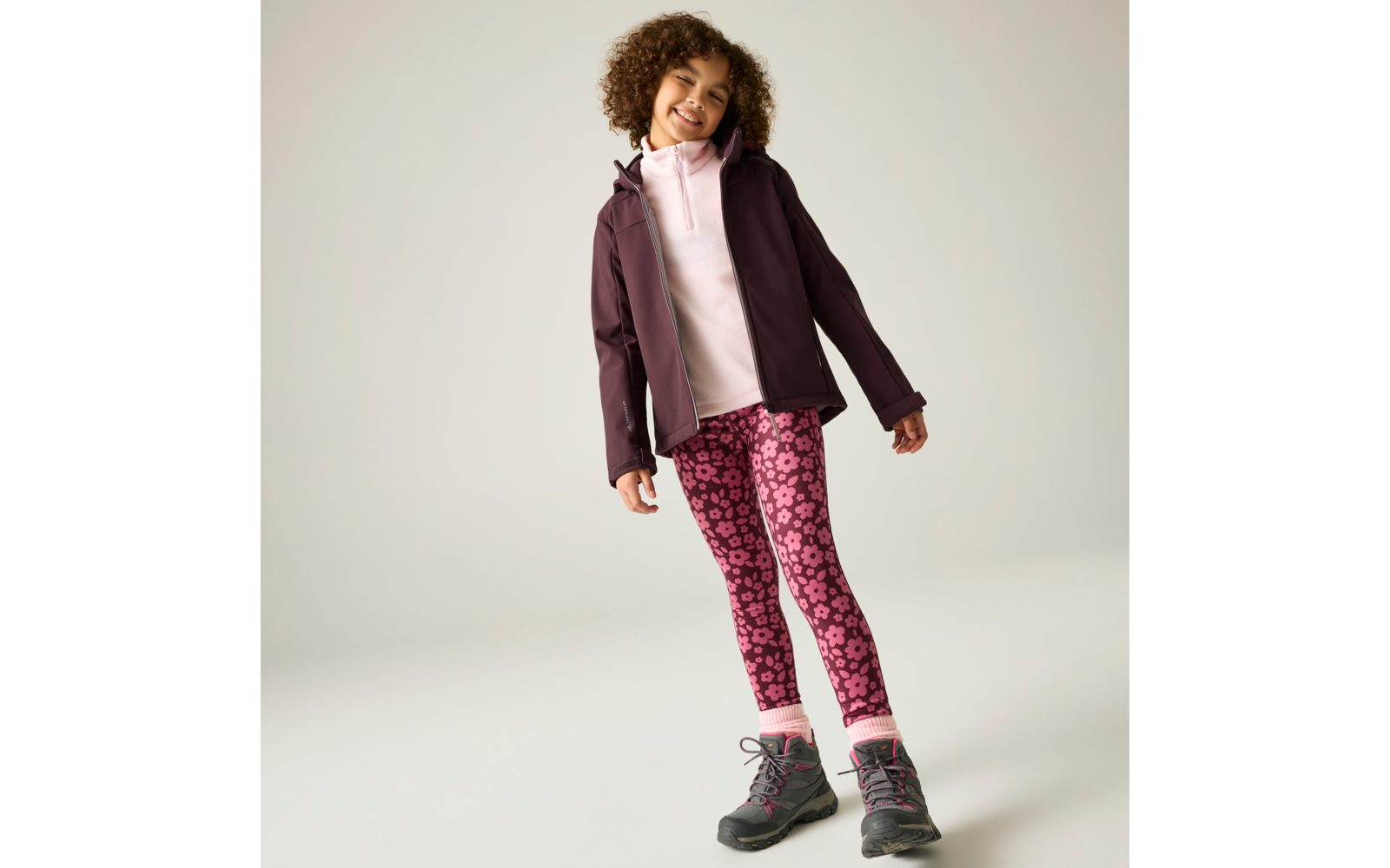 Kind mit lockigem Haar steht leicht geneigt, trägt eine dunkelviolette Jacke, einen hellrosa Kapuzenpullover, pinke Leggings mit Blumenmuster und graue Wanderschuhe mit rosafarbenen Details vor hellem Hintergrund