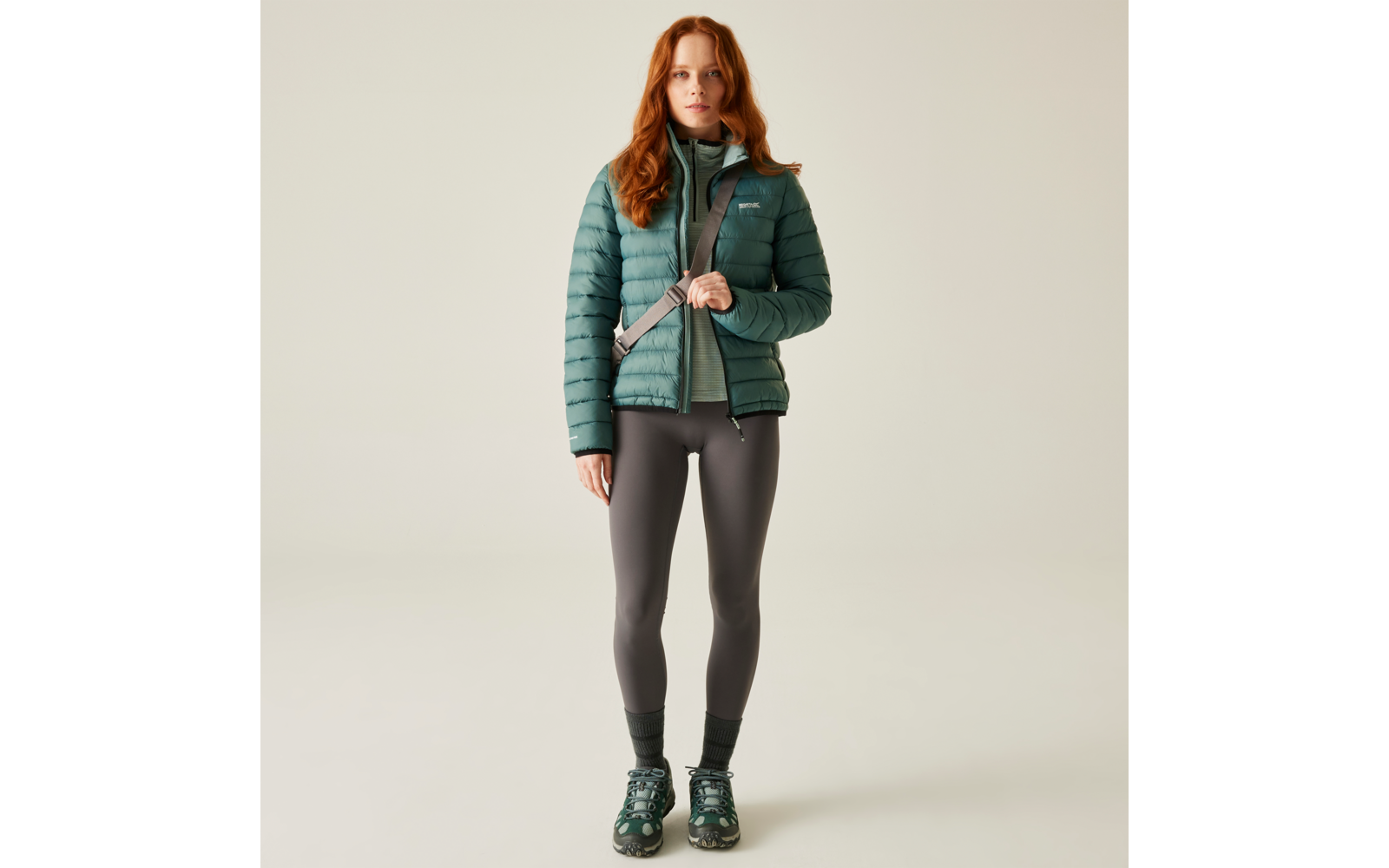 Frau mit rotem Haar trägt grüne gesteppte Jacke, graues Oberteil, graue Leggings und grüne Schuhe, vor hellem Hintergrund